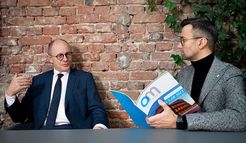 Dwaj elegancko ubrani mężczyźni siedzący przy szarym stole, rozmawiają, jeden z nich trzyma w ręku egzemplarz QMJ