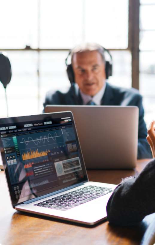 Otwarty laptop leżący na stole, w tle mężczyzna z słuchawkami na głowie siedzacy przed drugim otwartym laptopem