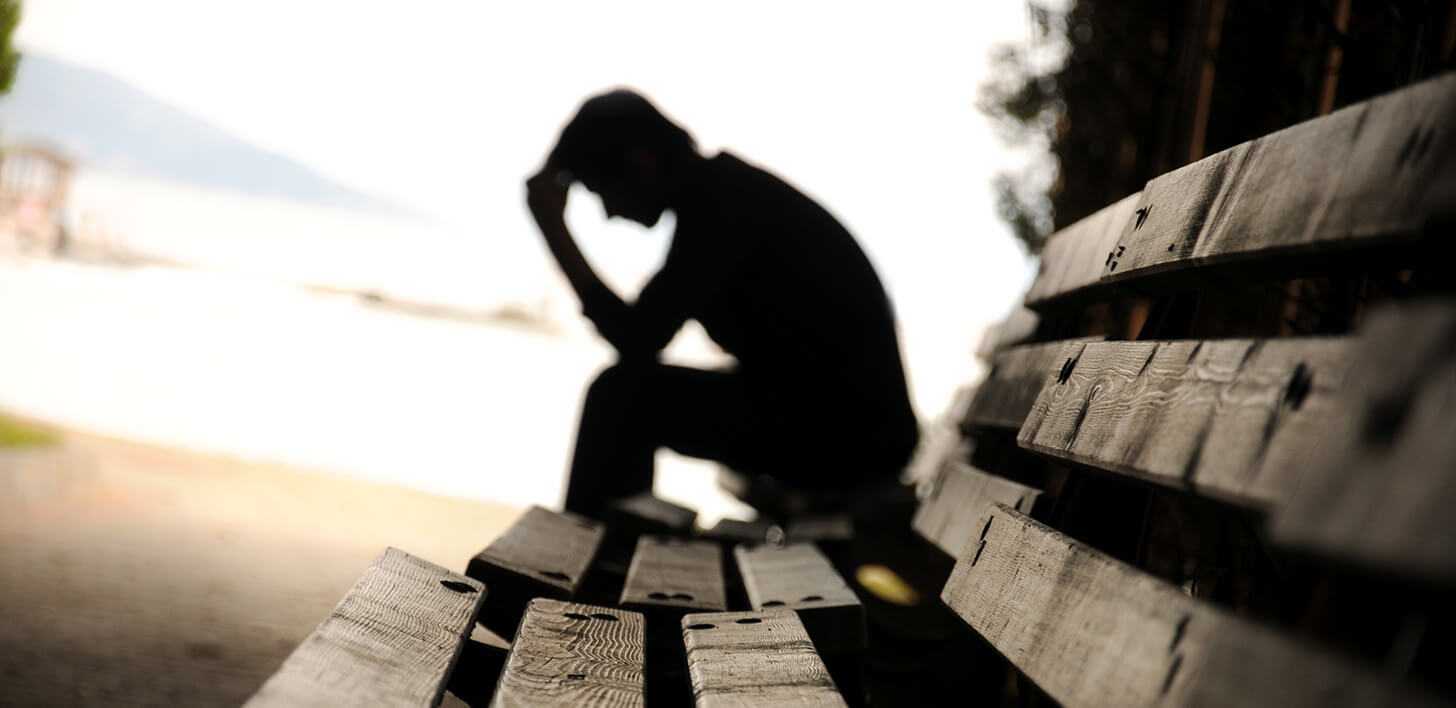 Czarna sylwetka postaci, siedząca na ławce, widziana z profilu, podpierająca głowę